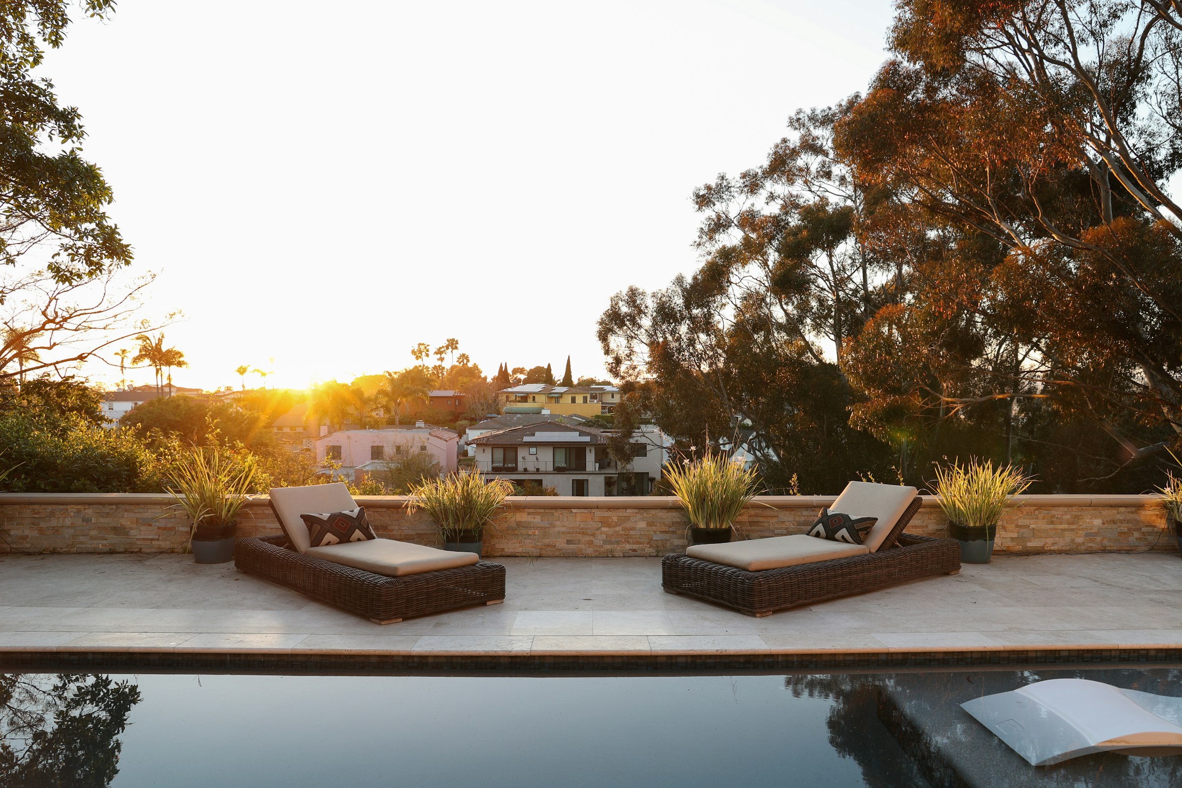 Stone pool deck with two loungers at golden hour, trees and rooftops beyond